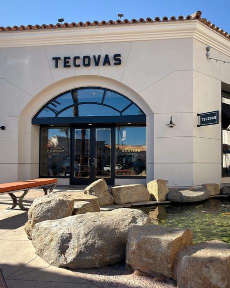A Tecovas storefront with arched glass doors, a rock-lined pond in front, a wooden bench, and clear sunny skies.