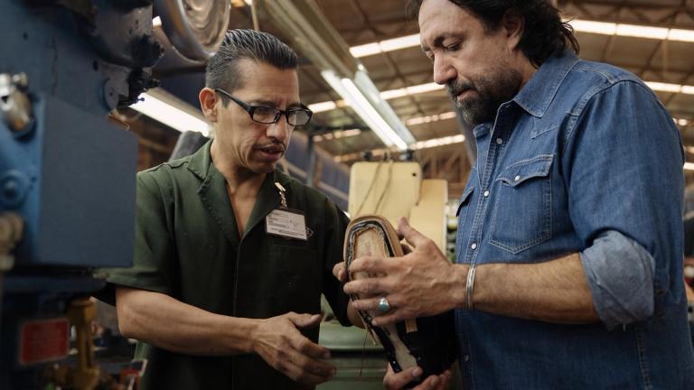 Two men stand in a factory, closely inspecting a shoe; one wears glasses and a name badge, while the other wears a denim shirt.