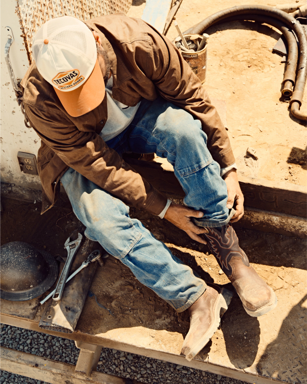 LongHaul Waterproof Square Composite Toe Work Boot in Tobacco / Black Cowhide. A man in a brown jacket, blue jeans, and a trucker hat sits on a step and adjusts his LongHaul Waterproof Square Composite Toe Work Boot Cowhide; work tools and equipment are scattered nearby on the ground.