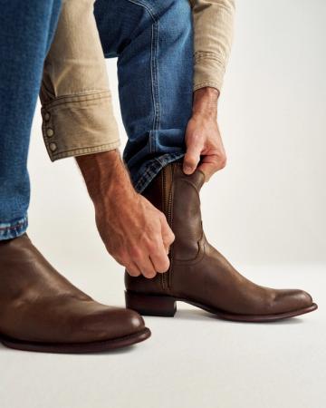 Person putting on brown leather cowboy boots, wearing blue jeans and a beige shirt.