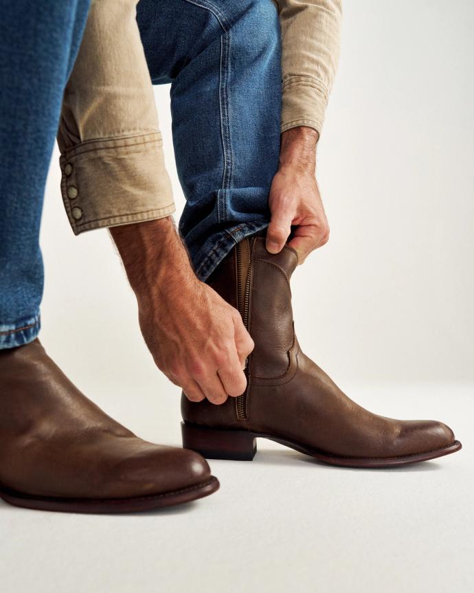 Person putting on brown leather cowboy boots, wearing blue jeans and a beige shirt.