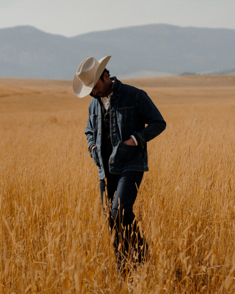 A person wearing a cowboy hat and denim jacket walks through a field of tall, dry grass.