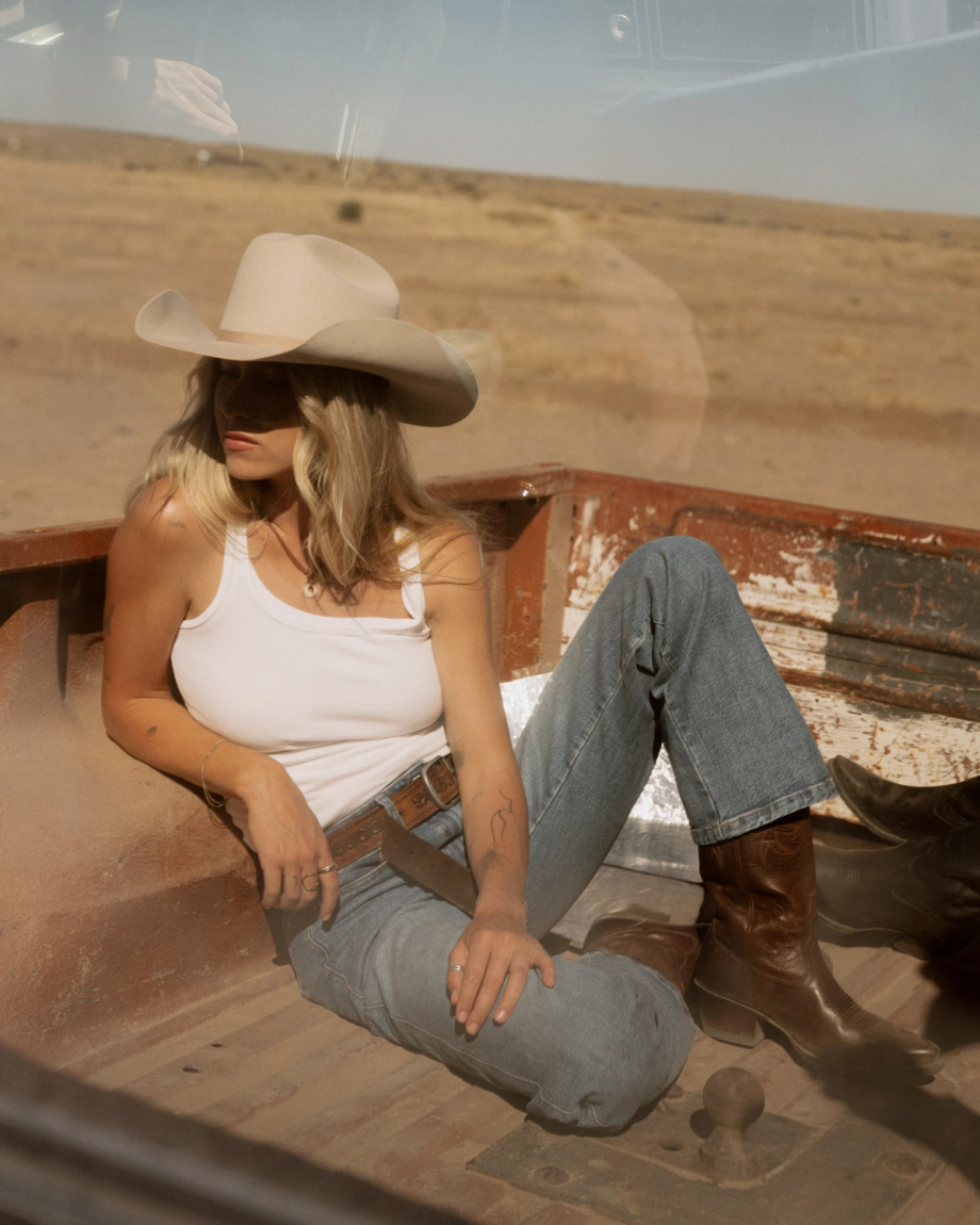 The Annie in Sequoia Cowhide. A woman in a cowboy hat, white tank top, jeans, and boots sits in the bed of a rusty pickup truck, her Annie Cowhide bag resting beside her in the dry, open landscape.