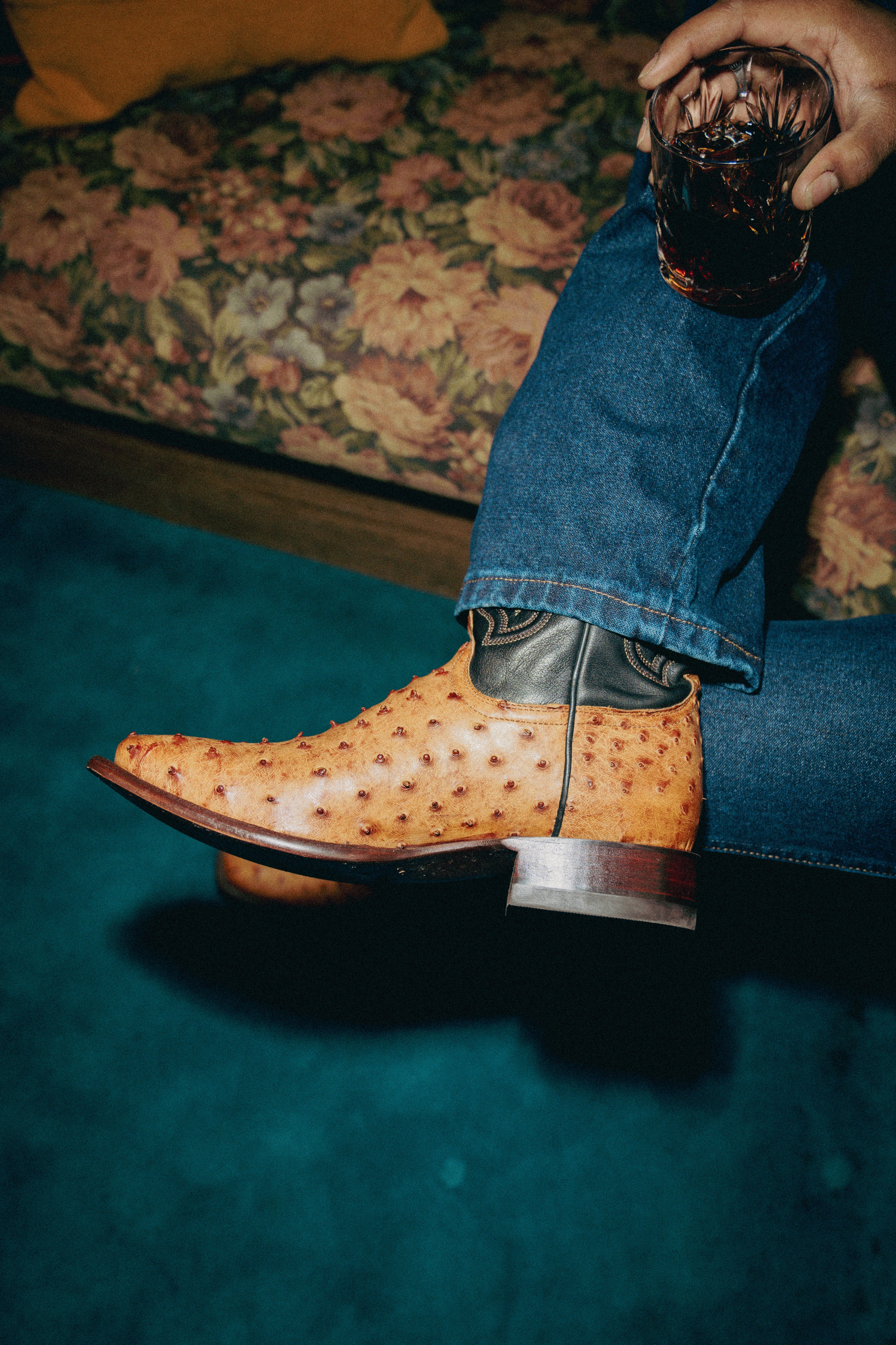 A person wearing jeans and tan ostrich leather cowboy boots sits on a floral couch with their legs crossed, holding a glass of dark beverage.