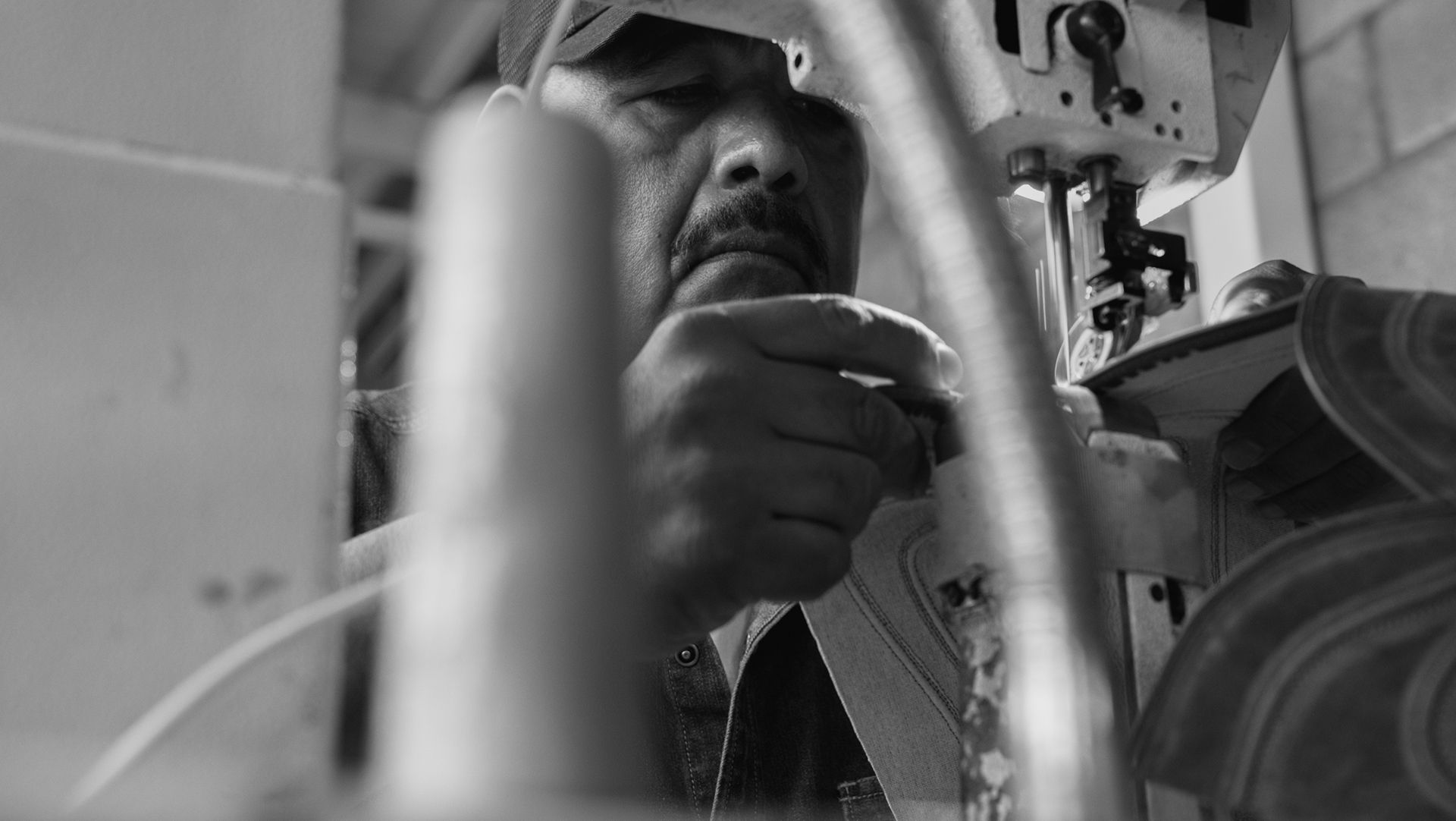 A person operates an industrial sewing machine, closely guiding material through the needle in a workshop setting.