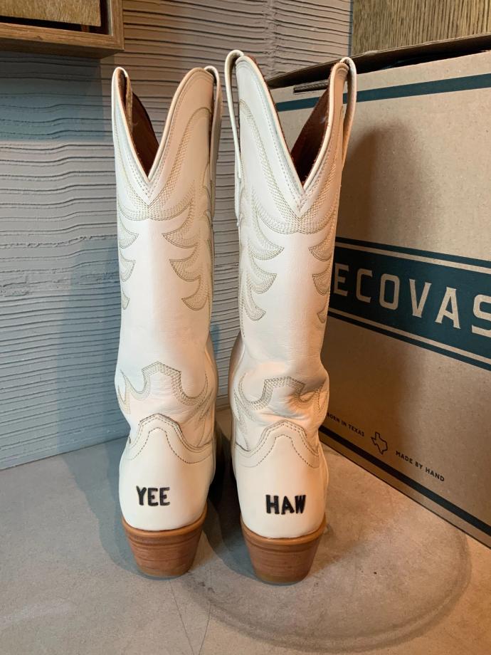 A pair of white cowboy boots with embossed patterns, displaying the words "YEE" on the left boot and "HAW" on the right boot. A cardboard box labeled "TECOVAS" is in the background.