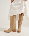 A person wearing a long white dress lifts the hem to reveal tan suede cowboy boots with decorative stitching.