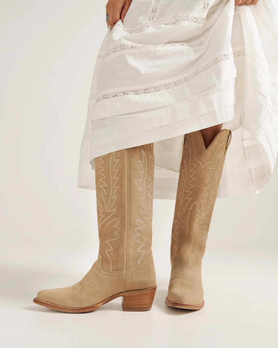 A person wearing a long white dress lifts the hem to reveal tan suede cowboy boots with decorative stitching.