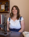A smiling woman with long hair sits at a table with a sewing machine, wearing a white top and denim overalls. There are rolls of fabric and framed pictures in the background.