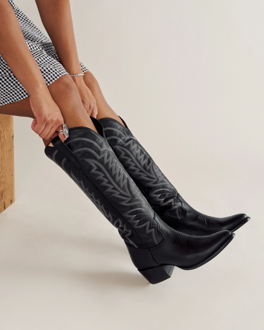 A person wearing a black-and-white checkered skirt is putting on tall black cowboy boots with decorative stitching.