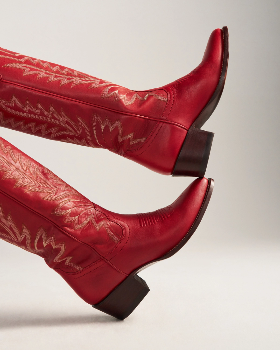 The Abby in Crimson Cowhide. Two legs wearing Crimson Cowhide red leather cowboy boots with decorative stitching are raised against a neutral background.
