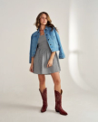 A person in a denim jacket, checkered dress, and red cowboy boots stands on a plain background.