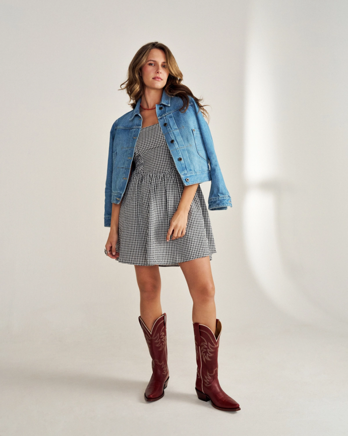 A person in a denim jacket, checkered dress, and red cowboy boots stands on a plain background.