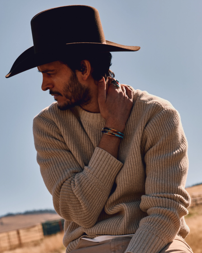 A person in a brown cowboy hat and beige sweater touches their neck while outdoors, wearing several bracelets.