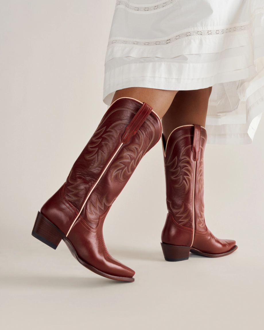 A person wearing a white dress and brown leather cowboy boots with decorative stitching and a low heel, standing against a plain background.