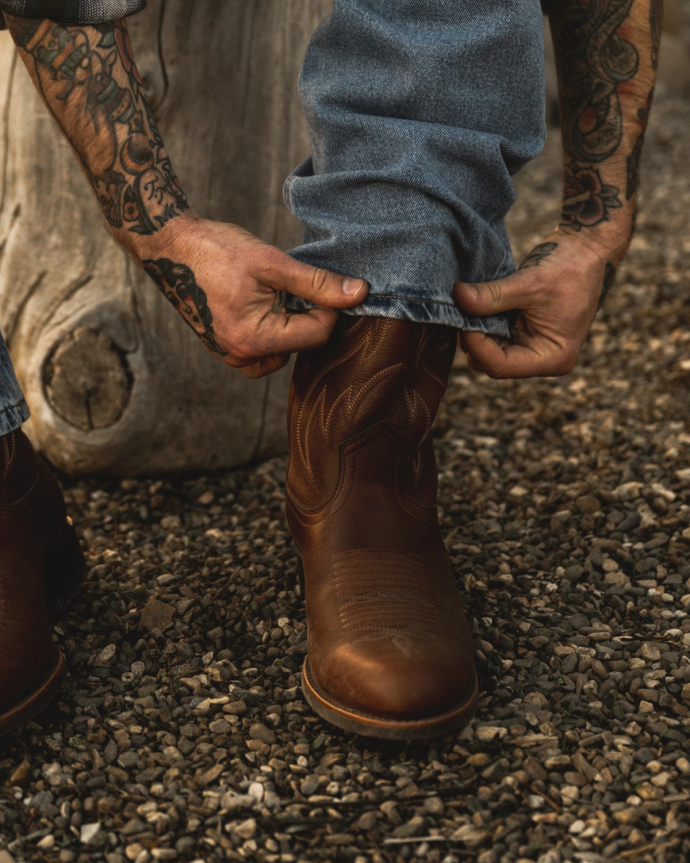 A person with tattooed arms adjusts the cuff of blue jeans over brown cowboy boots while standing on gravel.