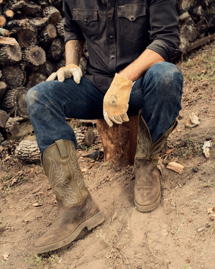 A person wearing gloves, jeans, and The Rugged Round Toe Cowhide boots in Umber / Sage sits on a tree stump outdoors, with a stack of chopped wood in the background.
