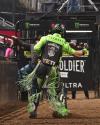 A rodeo rider in green gear and chaps stands in a dirt arena near a gate, with sponsor logos visible in the background.