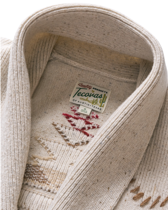 Close-up of a cream-colored knitted sweater with a collar, featuring a label that reads "Quality Products Tecovas of Austin, Texas, USA" and patterned designs in red and beige.