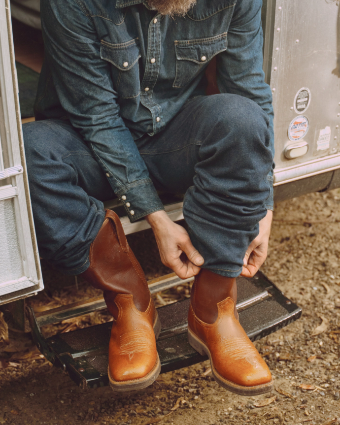 Person in denim outfit sitting on a step, putting on brown leather boots.