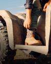 A person in jeans and cowboy boots sits on an ATV, adjusting their boot with one hand under sunlight.
