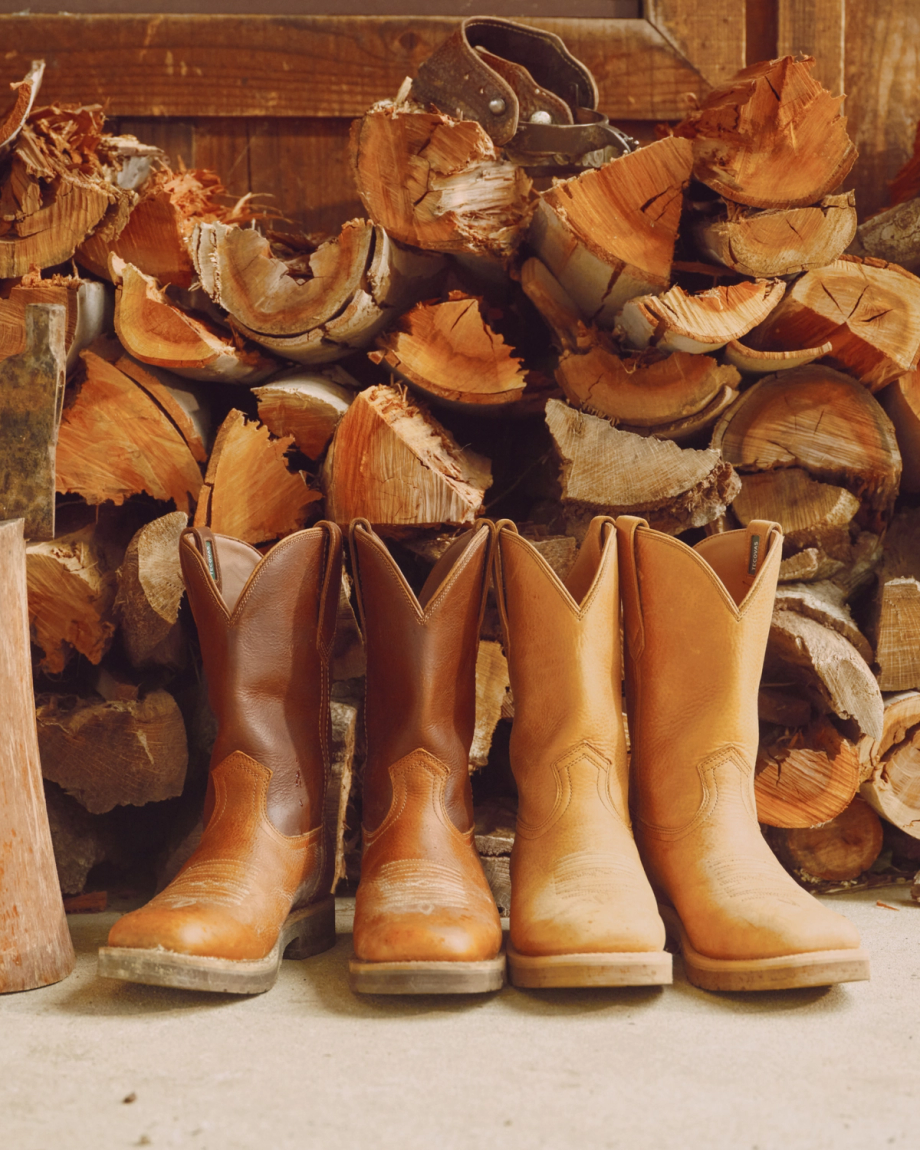 Four pairs of cowboy boots lined up in front of stacked firewood.