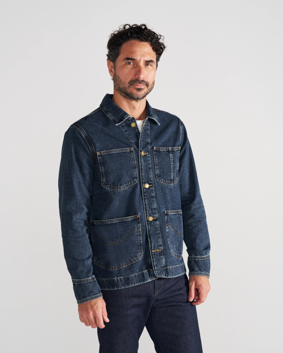 A man with short curly hair and a beard wears a denim jacket over a t-shirt and jeans, standing against a plain white background.