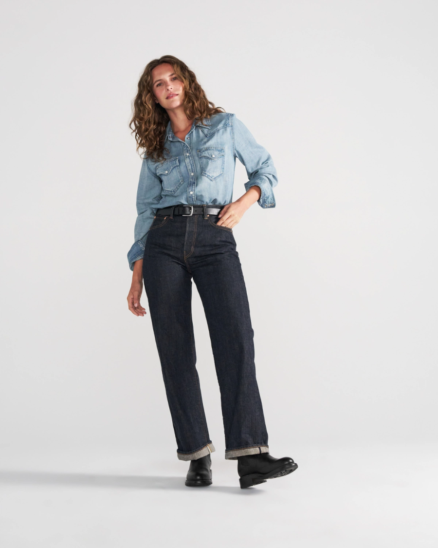 The Chelsea in Midnight Cowhide. A person stands against a plain background wearing a light denim shirt, dark cuffed jeans, a black belt, and sleek black boots crafted from midnight cowhide.