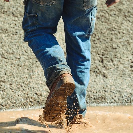 A person wearing blue jeans and brown boots steps into a muddy puddle, splashing water and dirt.