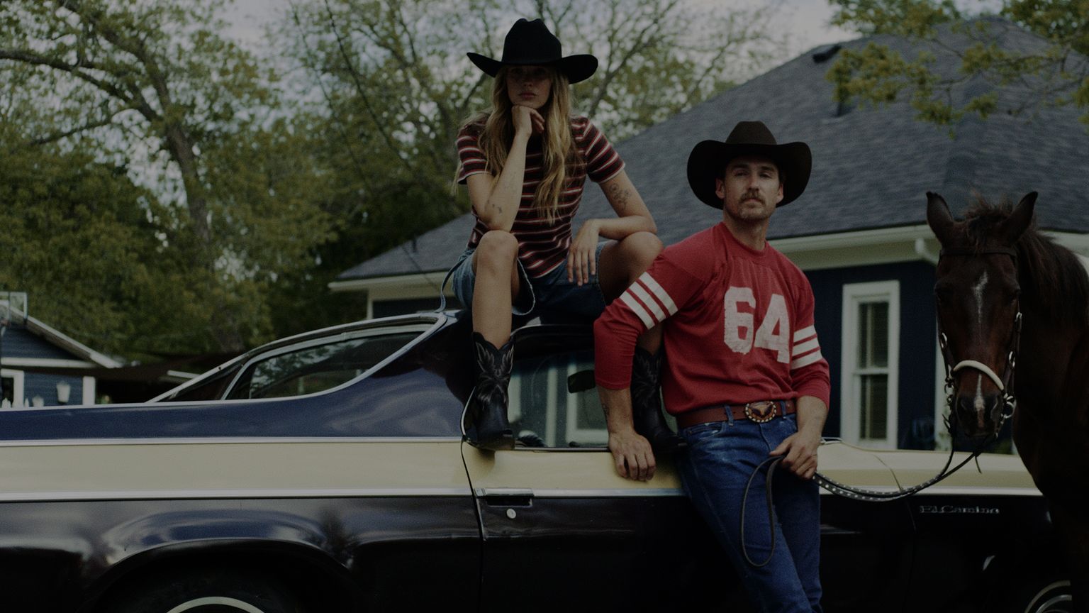 A woman and a man in cowboy hats pose by a vintage car with a horse nearby, in front of a house with trees in the background.