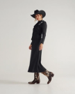 The Sadie in Hair on Hide Hair on Hide / Cowhide. Woman in a black cowboy hat, black jacket, black pleated skirt, and patterned Hair on Hide / Cowhide boots, standing and posing against a plain light background.