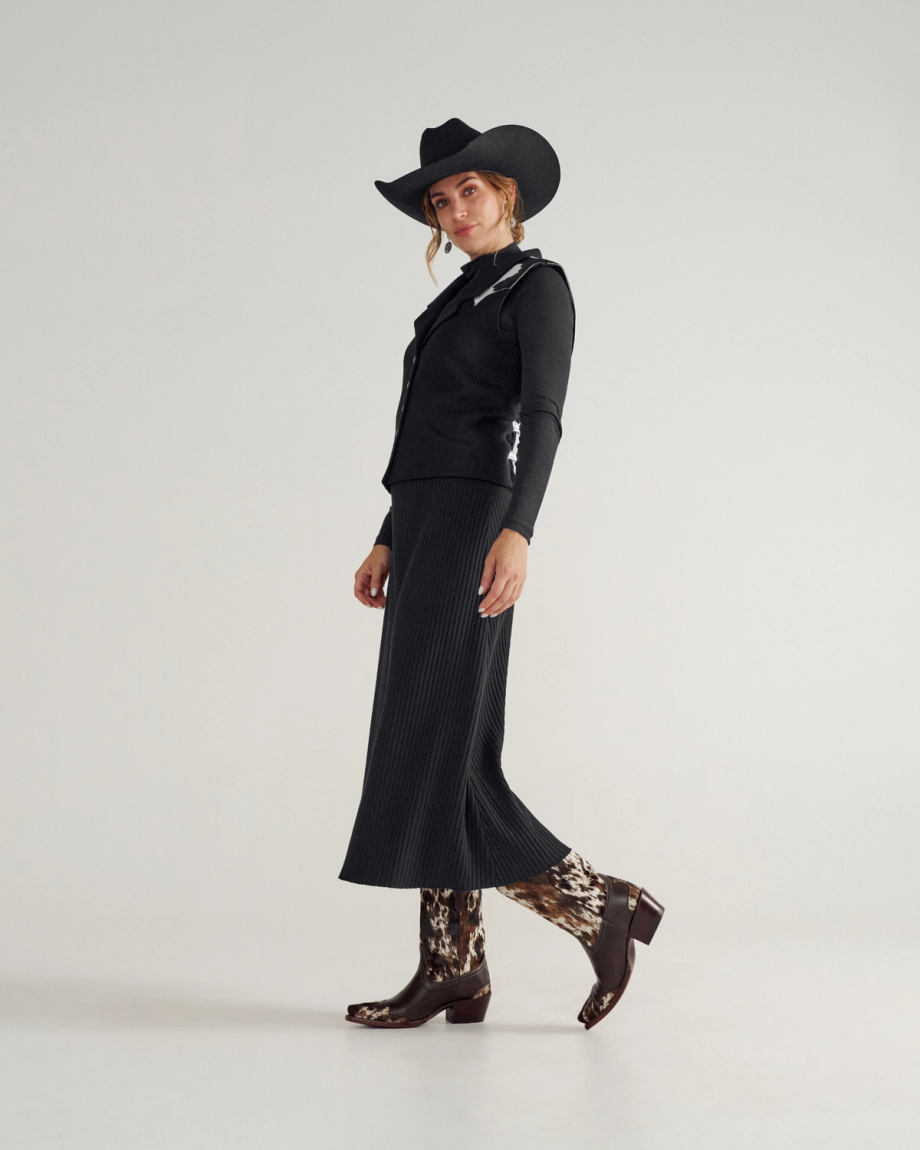The Sadie in Hair on Hide Hair on Hide / Cowhide. Woman in a black cowboy hat, black jacket, black pleated skirt, and patterned Hair on Hide / Cowhide boots, standing and posing against a plain light background.