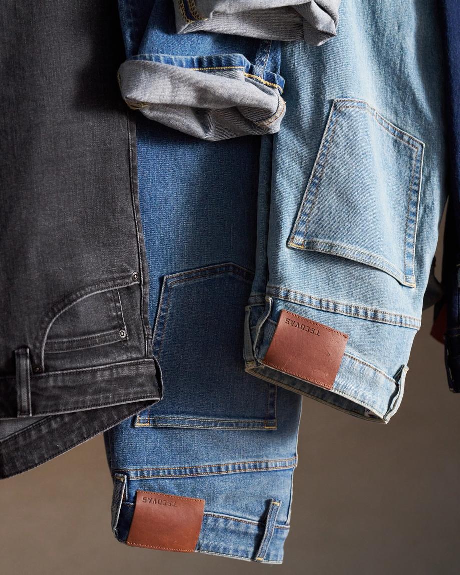 Three pairs of jeans, including black, medium blue, and light blue, are hung side by side with visible brown leather patches and rolled-up cuffs.