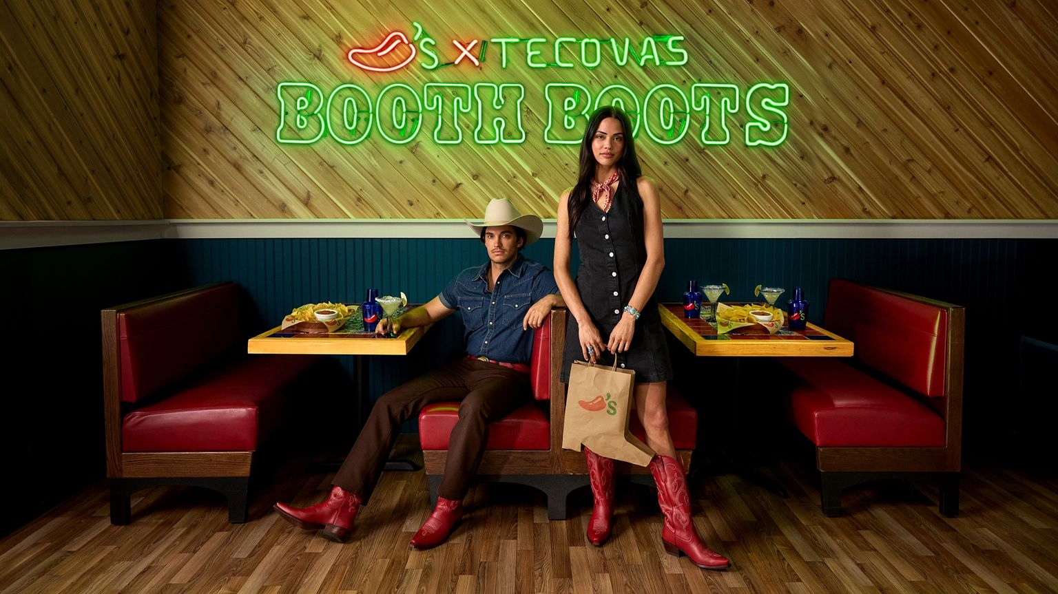 A man and woman wearing cowboy hats and red boots stand in a restaurant booth with red seats, under a neon sign that reads "Tecovas Booth Boots.