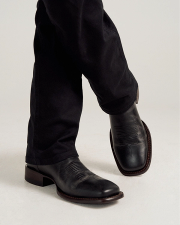 Person wearing black leather cowboy boots and black pants, standing with legs crossed on a light-colored surface.