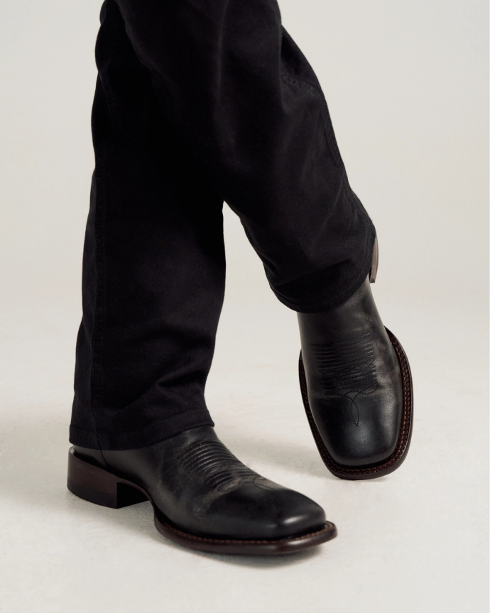 Person wearing black leather cowboy boots and black pants, standing with legs crossed on a light-colored surface.