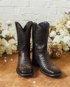 A pair of black leather cowboy boots with alligator skin texture, standing on a brown surface against a backdrop of white flowers.
