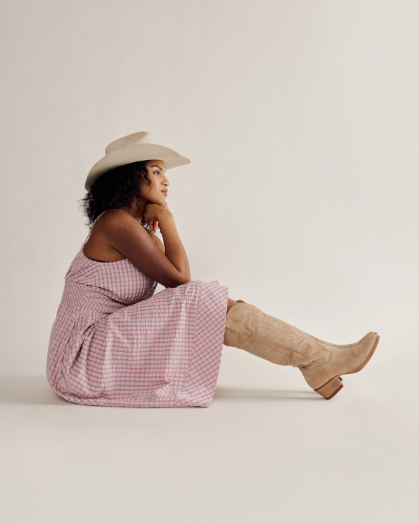 A woman in a pink plaid dress, tan cowboy boots, and a white cowboy hat sits on the floor facing sideways against a plain background.