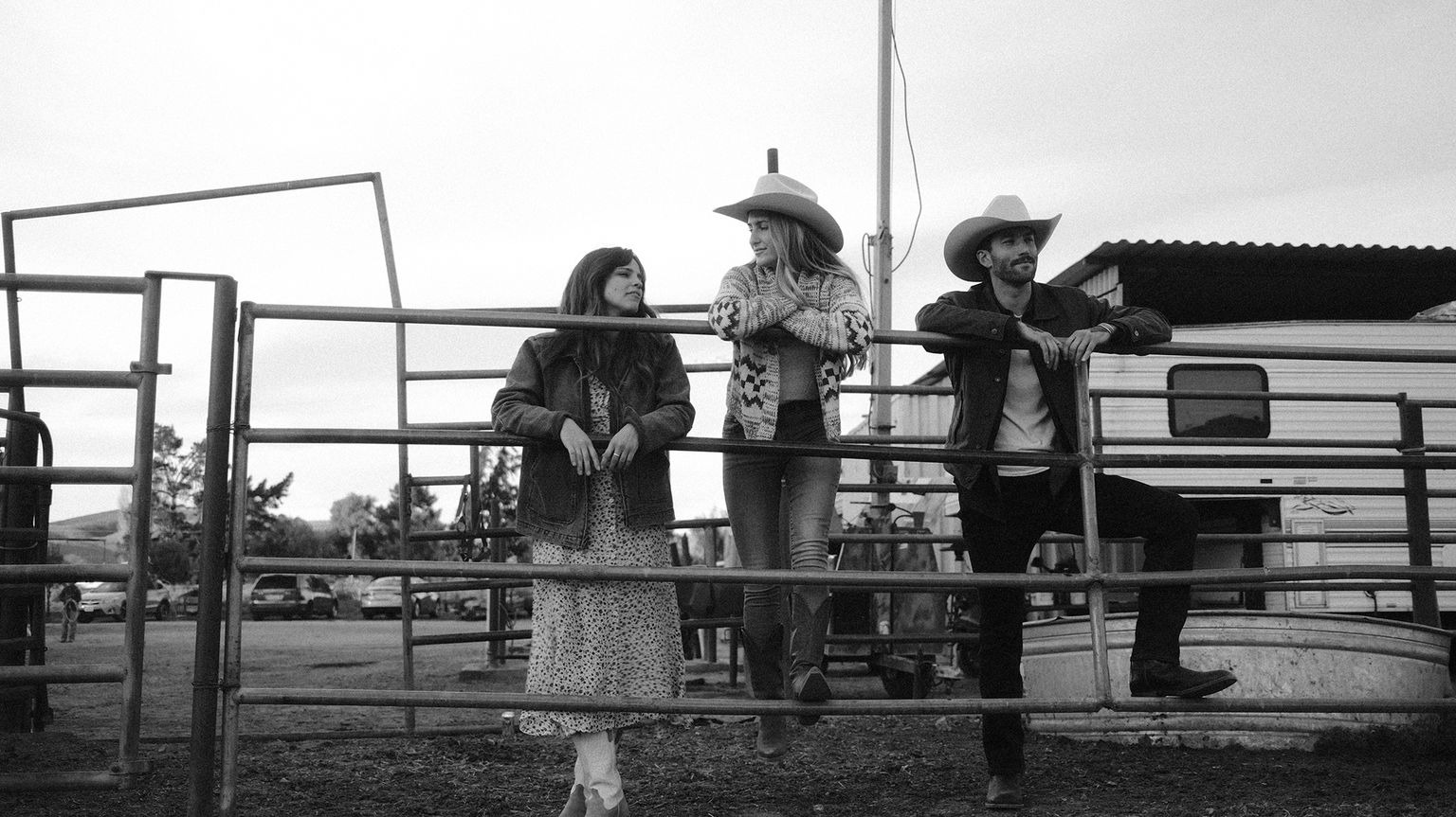 Three people in Western attire stand leaning on a metal fence in a rural setting, with trailers and equipment visible in the background.