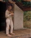 A woman stands on a wooden porch holding a cup. She is dressed in a beige sweater, white pants, and slippers, with a tent and greenery in the background.