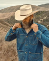 A bearded man adjusts his denim jacket while wearing a cowboy hat in a hilly, dry landscape.