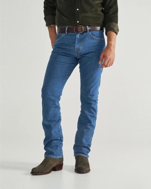 A person wearing a white short-sleeve shirt, blue jeans, and brown boots stands with one hand in their pocket against a plain background.