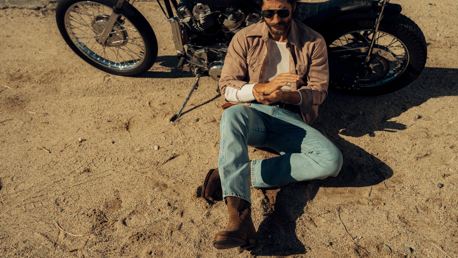 A man wearing sunglasses and boots sits on sandy ground next to a motorcycle, rolling up his sleeve.