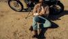 A man wearing sunglasses and boots sits on sandy ground next to a motorcycle, rolling up his sleeve.