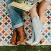 Two people sitting on a quilt with a guitar and vinyl records, showcasing their boots.