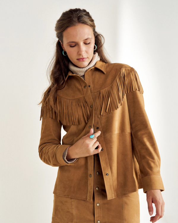A person wearing a tan Suede fringe jacket over a turtleneck sweater looks down with a neutral expression, adjusting the jacket.