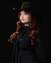 A woman with long, wavy auburn hair wears a black cowboy hat and black clothing, posing against a dark background.