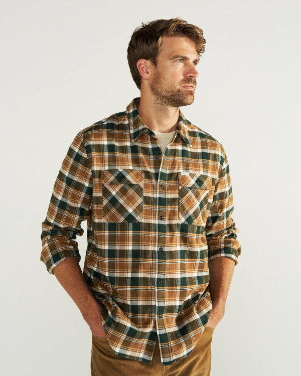 Man wearing a brown, green, and white plaid button-up shirt over a light t-shirt, standing with hands in pockets against a plain light background.