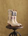 A pair of cream-colored cowboy boots with purple and blue floral embroidery sit on a rustic wooden stool against a tan textured wall.
