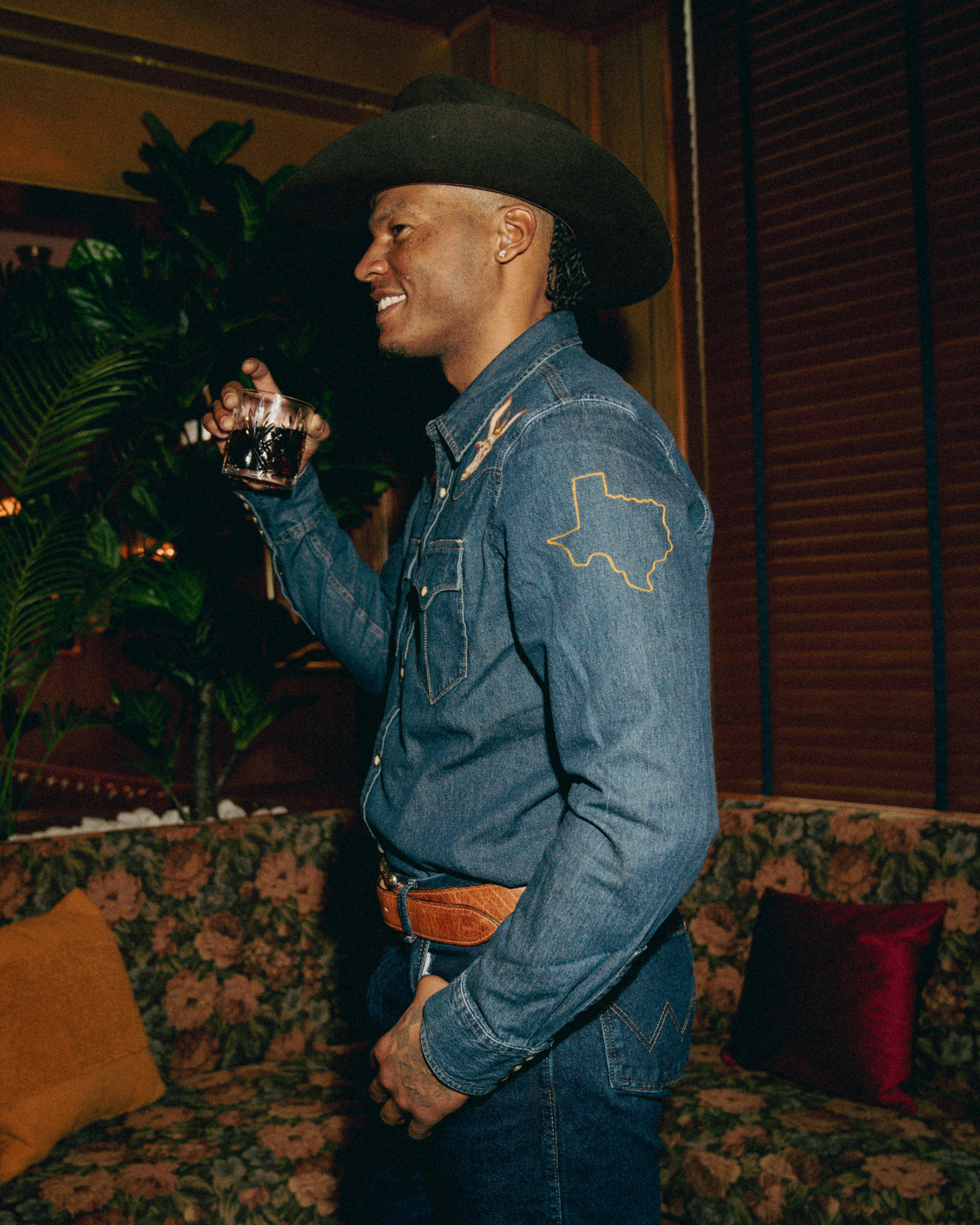 A man in a cowboy hat and denim outfit holds a drink, standing in a room with floral furniture and plants.